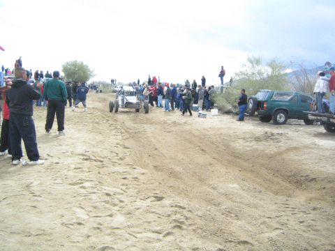 1990 Ford Bronco - off-road buggy drives along a sandy desert race course as spectators and parked vehicles line both sides.