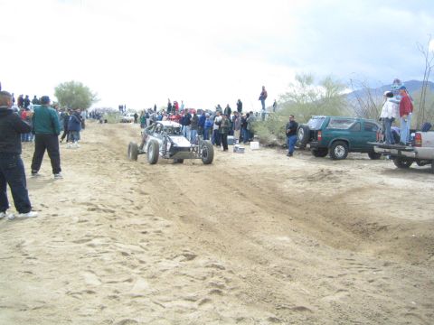 1990 Ford Bronco - silver tube-frame off-road buggy moves along a sandy desert track lined with spectators and parked pickups and SUVs.