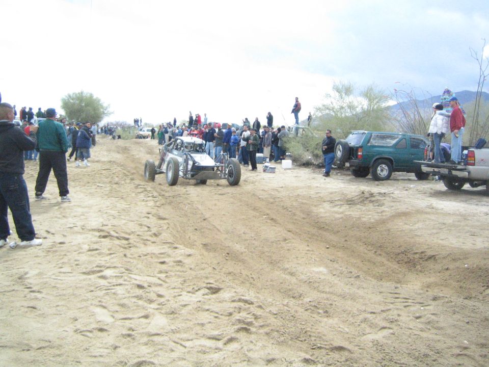 Silver tube-frame off-road buggy on sandy track with crowd and parked trucks