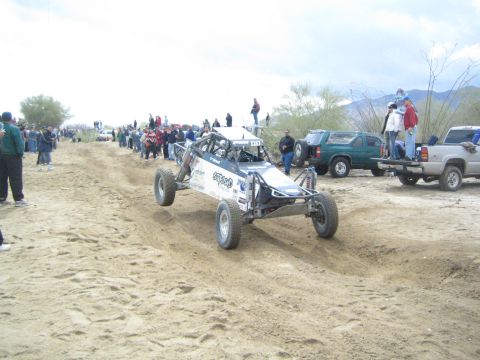 1990 Ford Bronco - white and blue tube-frame race buggy navigates a sandy desert course with spectators and parked trucks along the route.