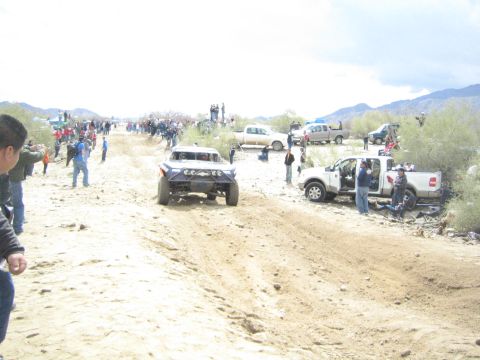 1990 Ford Bronco - dark off-road trophy truck runs a sandy desert course while a white pickup and spectators stand at trackside under a hazy sky.