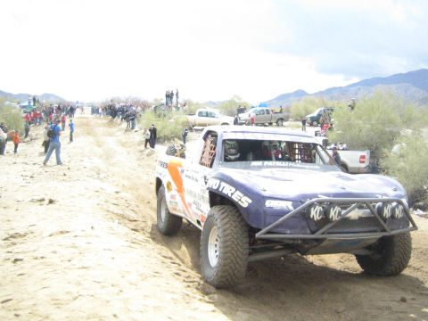1990 Ford Bronco - purple off-road trophy truck with KC light decals and sponsor markings drives a sandy desert track past spectators and parked pickups.