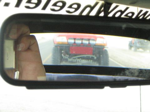 1990 Ford Bronco - rearview mirror reflects a lifted red truck with a roof light bar following close behind on a highway, with mirror text overhead.