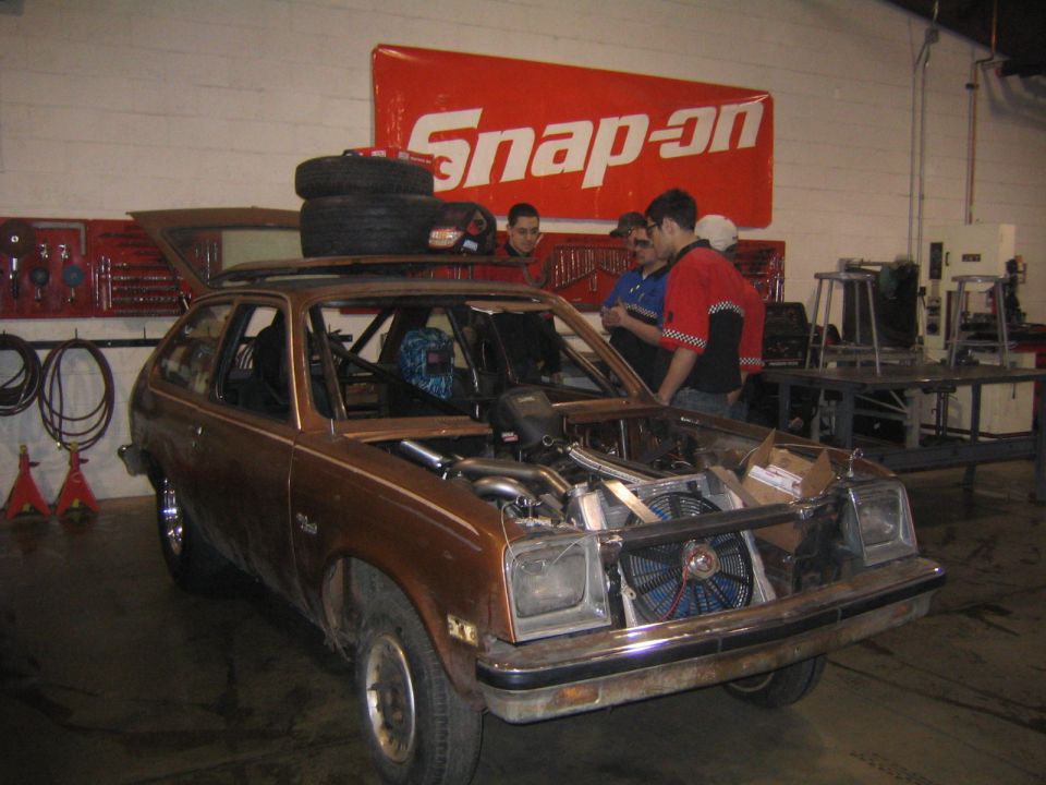 Brown hatchback car with hood open, exposed engine, and roll cage, in a garage