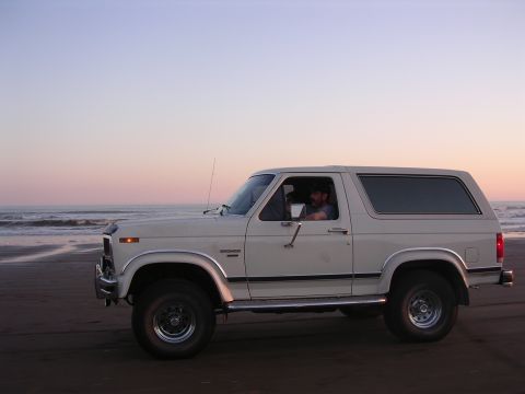 1986 Ford Bronco - A white 1986 Ford Bronco sits on the sand near the ocean as the sun sets.