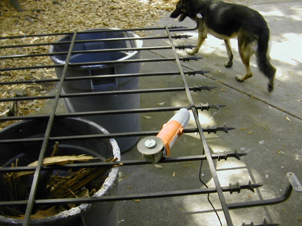 Wrought-iron fence section on trash cans with orange angle grinder, dog walking past