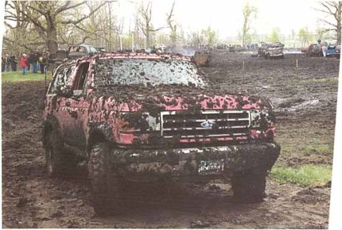 1993 Ford Taurus - A red SUV is heavily caked in mud, participating in a muddy event with other vehicles in the background.