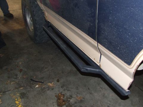1986 Ford Bronco - Dark blue SUV with custom black rock sliders and tan pinstriping visible from the side.