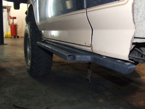 1986 Ford Bronco - Side view of a tan and dark blue Ford Bronco with custom rock sliders and large tires, lifted in a garage.