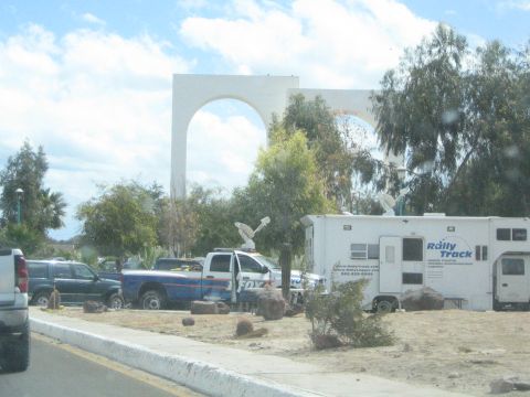 1990 Ford Bronco - large white arch monument rises behind parked trucks and a white Rally Track motorhome along a dusty roadside.