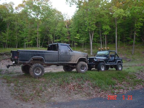 1984 Ford F-250 - Two off-road vehicles, a lifted pickup truck and a Jeep, are parked side-by-side on a grassy area next to a dirt path.