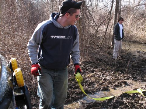 1995 Ford Bronco - Two people prepare to use a tow strap to pull a vehicle out of a muddy, wooded area.
