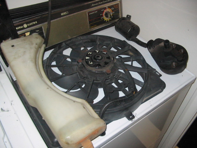 Radiator fan and plastic coolant reservoir sitting on top of a washing machine. Also visible are two black cylindrical components