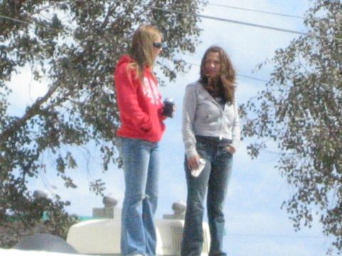 1990 Ford Bronco - Two young women, one in a red hoodie and one in a gray jacket, stand atop a light-colored vehicle roof framed by bare trees and blue sky.