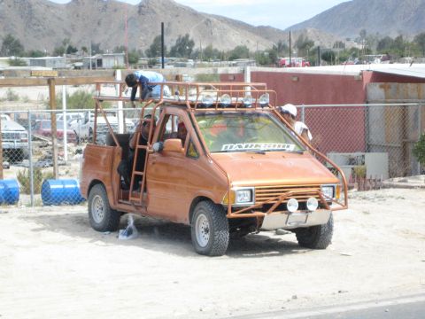 1990 Ford Bronco - orange custom off-road pickup with a tall roof rack and light pods is parked on sand near a chain-link fence with desert hills behind.