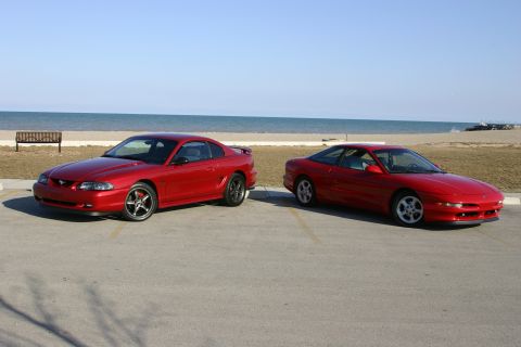 1993 Ford Probe - Two red coupes are parked in a lot with a beach and ocean in the background.
