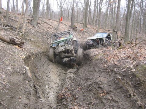 1992 Ford Bronco - Two off-road vehicles struggle through a deep, muddy trench on a wooded slope, with one vehicle partially submerged in mud.