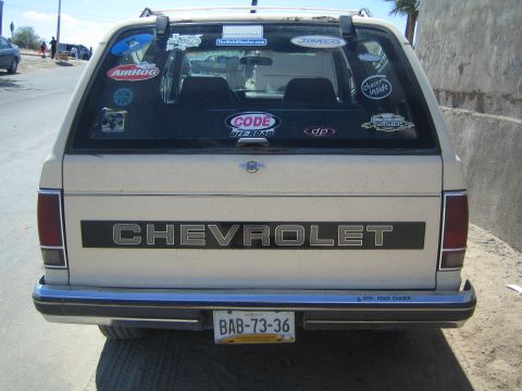 1990 Ford Bronco - tan rear of a sticker-covered Chevrolet SUV shows the CHEVROLET tailgate lettering and a yellow Mexican license plate along a roadside.