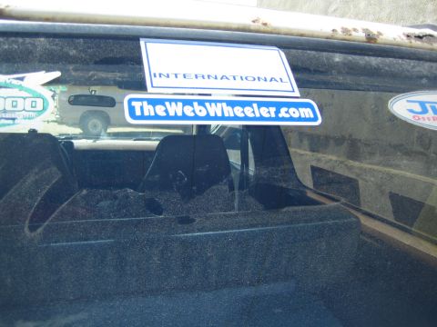 1990 Ford Bronco - close-up of a dusty rear window shows an INTERNATIONAL banner over a blue TheWebWheeler.com decal among other off-road stickers.