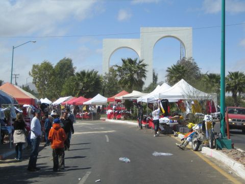 1990 Ford Bronco - Rows of white and red vendor tents line a street market beneath twin white arches, with people and a red pickup along the roadside.