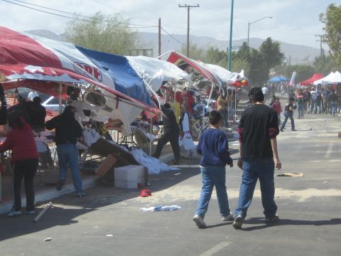 1990 Ford Bronco - Wind-toppled red and white vendor tents and scattered debris line a street as people walk past under a hazy sky with hills behind.