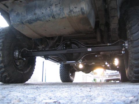 1990 Ford Bronco - Low ground-level shot of a lifted truck's undercarriage showing the rear solid axle, track bar, leaf springs and chunky mud-terrain tires.
