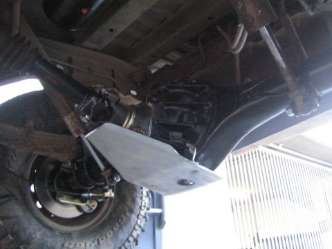 1990 Ford Bronco - Underbody view of a lifted 4x4 showing a metal skid plate, leaf-spring suspension, shock absorber and a mud-terrain tire.