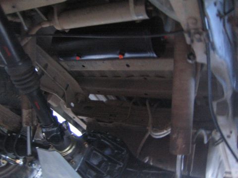 1990 Ford Bronco - Dim undercarriage shot showing a vehicle's driveshaft, fuel tank, frame rails and routed lines and cables.