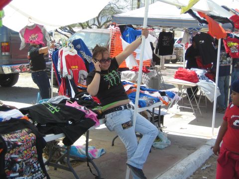 1990 Ford Bronco - person talks on a phone while seated among hanging graphic T-shirts and folding tables at an outdoor merchandise booth.