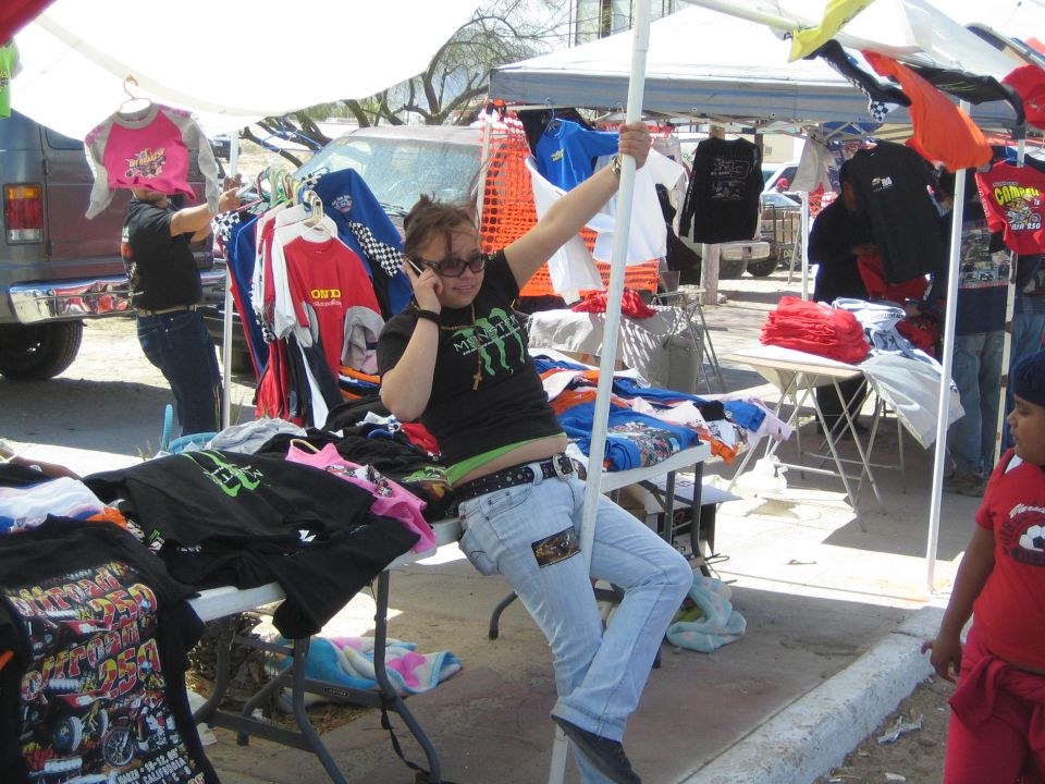 Person on a phone seated among racks of graphic T-shirts at an outdoor vendor booth
