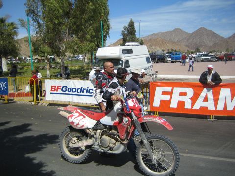 1990 Ford Bronco - Riders sit on a red-and-white Honda dirt bike marked 348 in a paved staging area with BFGoodrich and FRAM banners and desert mountains behi…