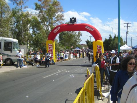 1990 Ford Bronco - red and yellow inflatable Tecate arch spans a wide street lined with crowds, yellow barriers and a motorhome.