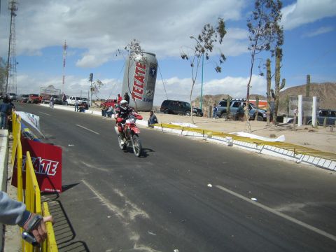 1990 Ford Bronco - helmeted rider on a red Honda dirt bike rides a desert road toward a giant inflatable Tecate beer can and parked vehicles.