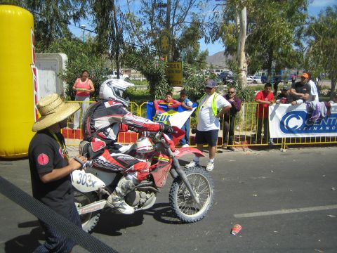 1990 Ford Bronco - person in a straw hat pushes a number 309x red Honda race dirt bike along a road past spectators behind barriers.
