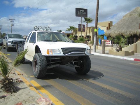 1990 Ford Bronco - white prerunner-style truck with a roof light bar and long-travel suspension parks on a street near a 'Bivalvo' sign and palm trees.