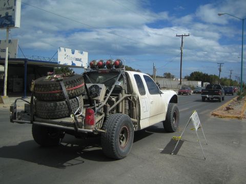 1990 Ford Bronco - mud-caked race prerunner truck with bed-mounted spare tires and roof spotlights drives a street past a roadside sign.
