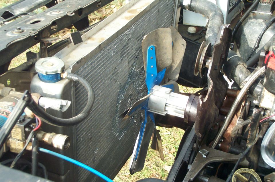 Close-up of a vehicle's radiator and fan assembly. Shows a blue fan with silver motor attached to a metal bracket