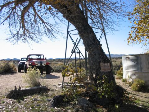 1992 Ford Bronco - Two off-road vehicles are parked in a dry, rural landscape under a large tree with metal supports.