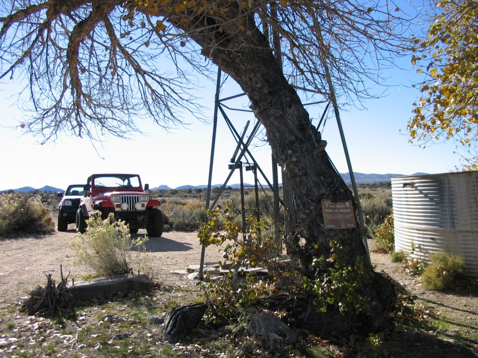 Red Jeep Wrangler and black SUV parked on dirt road near a large tree and metal water tank