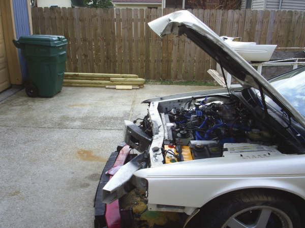 Car with hood open showing engine and some front end damage. Green trash can and wood in background