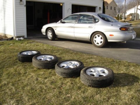 1999 Ford Taurus - A silver sedan is parked in front of a garage, with four tires laid out on the lawn.
