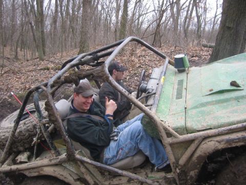 1992 Ford Bronco - Two people in a muddy off-road buggy appear stuck in a wooded area.