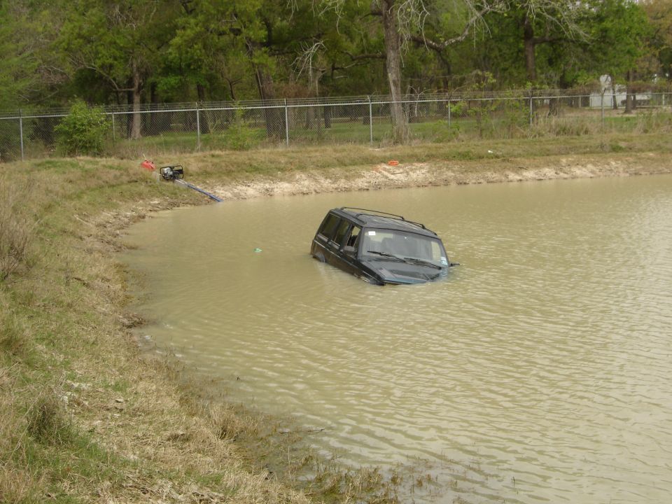 Black Jeep Cherokee partially submerged in murky water near a grassy bank with a pump and hose visible