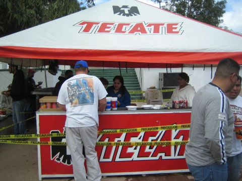 1990 Ford Bronco - red-and-white Tecate-branded concession booth strung with caution tape serves patrons under a peaked tent at an outdoor event.