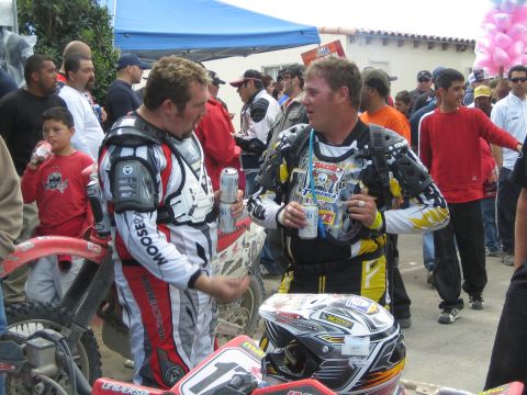 1990 Ford Bronco - Two dirt-bike riders in racing gear and chest protectors chat while holding drink cans amid a festive crowd.