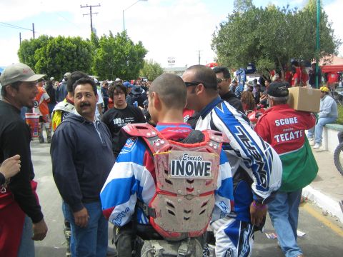 1990 Ford Bronco - Off-road riders in chest protectors stand in a crowd, one labeled Ernesto, near a green Tecate Score banner.