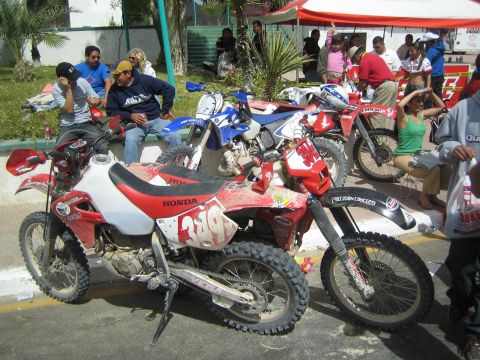 1990 Ford Bronco - red Honda dirt bike number 339 and other bikes are parked in a row on a street as spectators sit nearby.