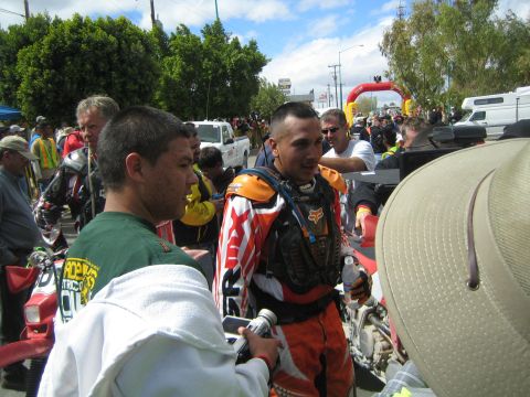 1990 Ford Bronco - motorcycle racer in orange Fox gear stands amid a crowd near an inflatable arch at an off-road race.