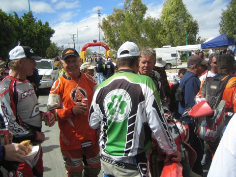 1990 Ford Bronco - Several off-road riders in racing jerseys chat and hold drinks near an inflatable arch and parked RVs.