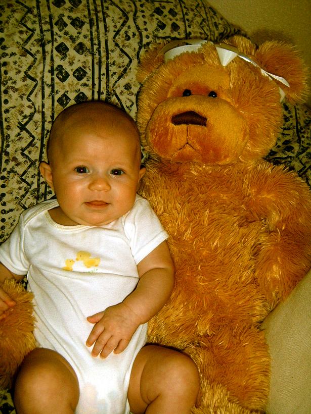 Baby sitting next to a large teddy bear on a patterned surface. Text at bottom reads SUPERMOTORS